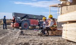 A small group of construction workers sit on a stack of lumber as they take a quick lunch break.  They are each wearing proper safety gear and are talking amongst themselves as they sit onsite.